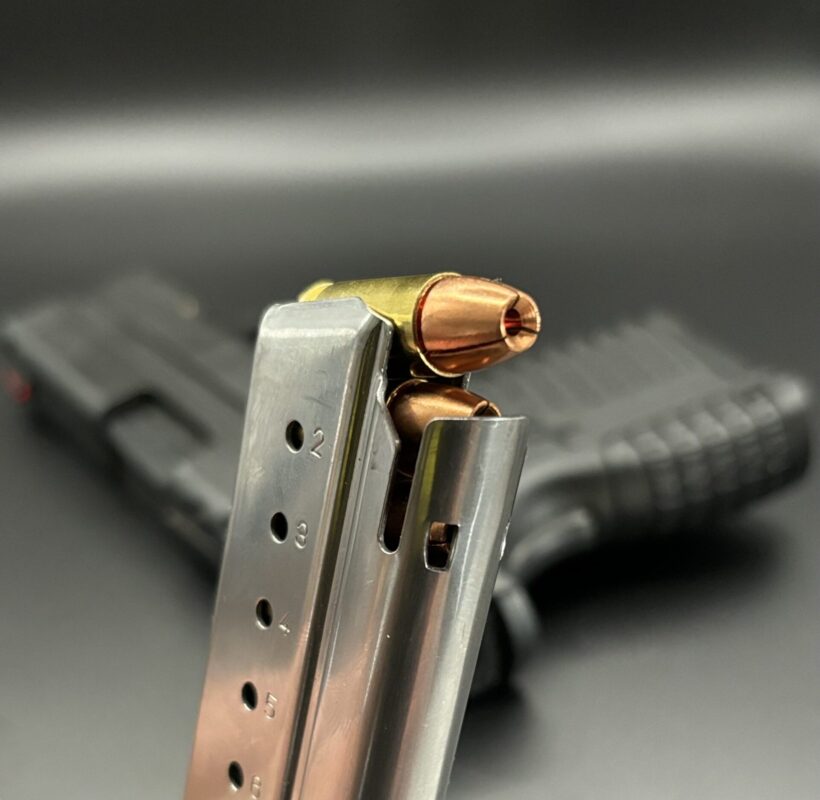 ms Ammunition – High-Quality Ammunition for Rifles and Shotguns" Close-up of a metal handgun magazine filled with high-quality ammo, featuring copper-colored bullets partially loaded. In the background, a blurred black handgun is visible. The focus is on the ammunition and magazine, emphasizing the metallic texture and intricate details of rifle cartridges and shotgun shells suitable for those seeking bulk ammunition. Bulk Ammo Available for Purchase"